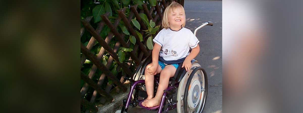 Die SMA-Patientin Amelie als Kind. Das Mädchen sitzt draußen im Rollstuhl und lächelt fröhlich.
