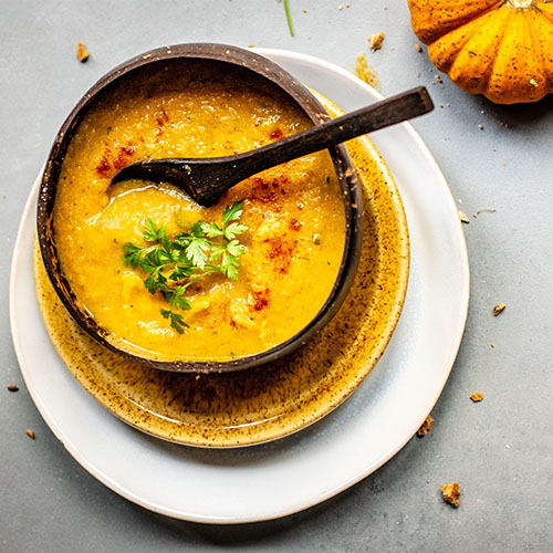 Eine orange Suppe oder püriertes Essen in einer Suppenschüssel, ein Esslöffel und Kürbisse sowie Kräuter.