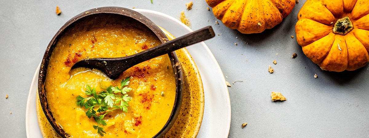 Eine orange Suppe oder püriertes Essen in einer Suppenschüssel, ein Esslöffel und Kürbisse sowie Kräuter.