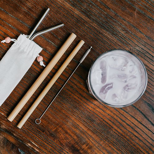 Wiederverwendbare Strohalme aus Holz und Metall eine Strohalmbürste zum Reinigen sowie ein Glas mit weißer Flüssigkeit und Eiswürfeln auf einem Holztisch.