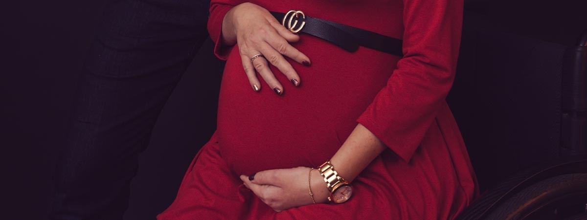 Der Bauch einer Schwangeren im Rollstuhl. Die Frau trägt ein rotes Kleid und hält ihren Bauch.