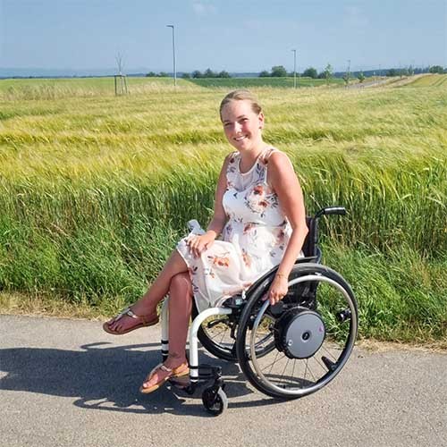 SMA-Patientin Svenja sitzt im Sommerkleid im Rollstuhl vor einem Feld.