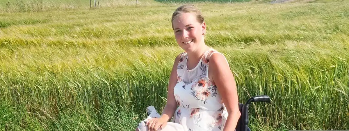 SMA-Patientin Svenja sitzt im Sommerkleid im Rollstuhl vor einem Feld.