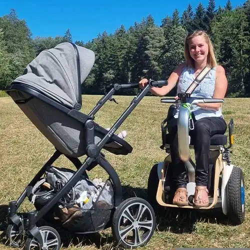 SMA-Patientin Svenja sitzt im Rollstuhl auf einer Wiese und schiebt einen Kinderwagen. Im Hintergrund sind ein Wald und blauer Himmel.