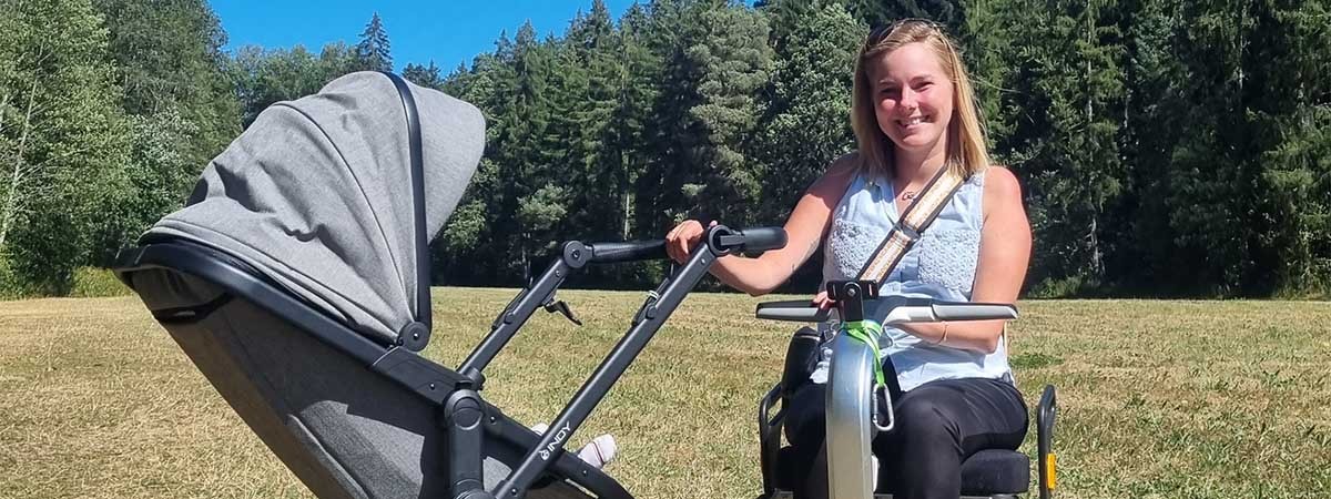 SMA-Patientin Svenja sitzt im Rollstuhl auf einer Wiese und schiebt einen Kinderwagen. Im Hintergrund sind ein Wald und blauer Himmel.