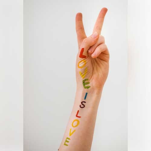 Auf einem Arm steht in bunten Regenbogen-Farben „LOVE IS LOVE“. Die Hand macht ein Peace-Zeichen.