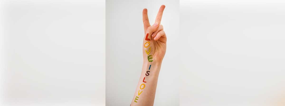 Auf einem Arm steht in bunten Regenbogen-Farben „LOVE IS LOVE“. Die Hand macht ein Peace-Zeichen.