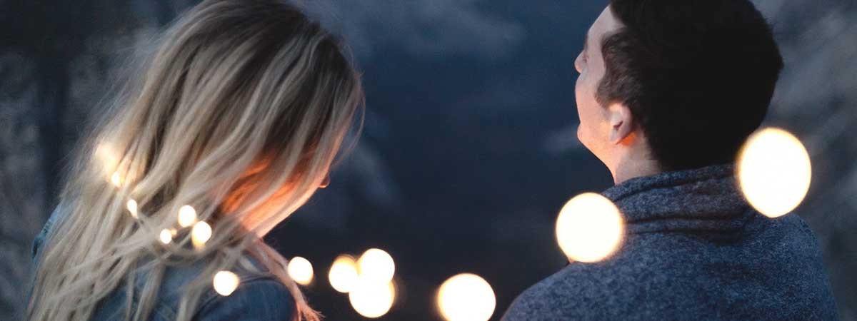 Eine Frau mit blonden Haaren und ein Mann mit dunklen Haaren sind von hinten zu sehen. Sie halten nachts Kerzen in den Händen und sind von Lichterketten umgeben, sodass eine romantische Dating-Stimmung entsteht. 
