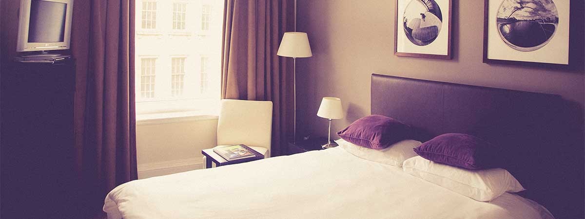 Ein Bett in einem Hotelzimmer in Braun- und Lila-Tönen und ein Fenster, durch das Licht hereinstrahlt, sind zu sehen.