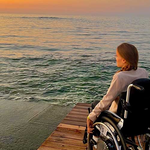 SMA-Betroffene Amelie sitzt in ihrem Rollstuhl auf einem Steg am Meer. Sie ist im Urlaub und genießt den Sonnenuntergang.