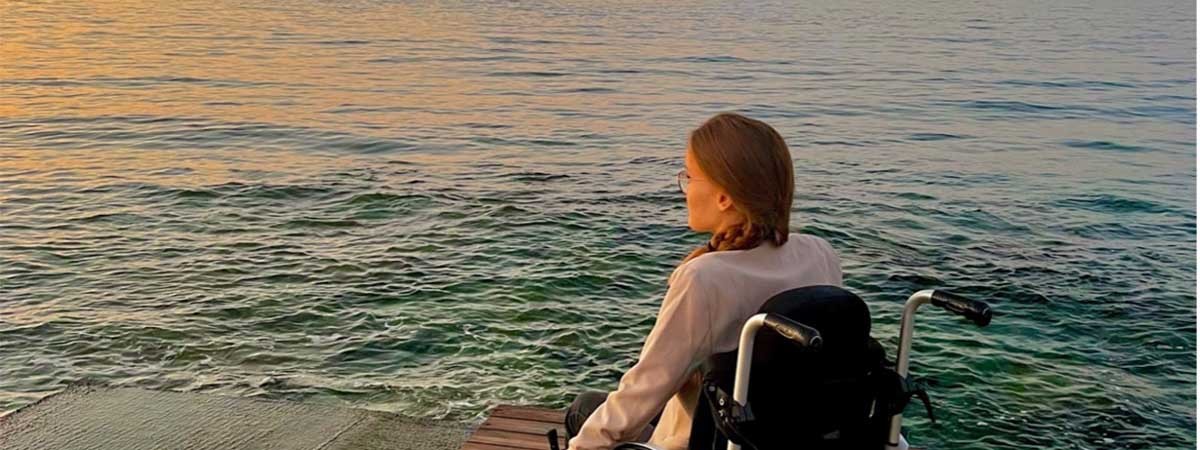 SMA-Betroffene Amelie sitzt in ihrem Rollstuhl auf einem Steg am Meer. Sie ist im Urlaub und genießt den Sonnenuntergang.