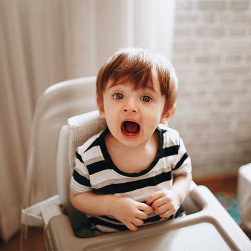 Ein Kleinkind sitzt mit aufgerissenem Mund in einem Kinderstuhl bzw. Hochstuhl für Kleinkinder.