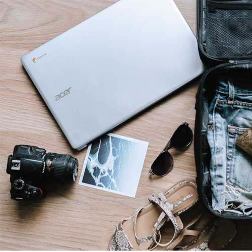 Ein Reisekoffer, der halb gepackt ist mit Kleidung für einen Urlaub und einem Kosmetikbeutel. Neben dem Reisegepäck liegen ein Laptop, eine Sonnenbrille, eine Kamera für Urlaubsbilder, ein Foto vom Meer und Sandalen.