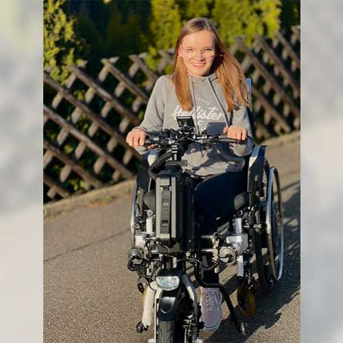 SMA-Betroffene Amelie fährt mit einem Zuggerät in ihrem Rollstuhl durch die Straßen und lächelt glücklich.