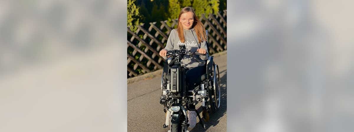 SMA-Betroffene Amelie fährt mit einem Zuggerät in ihrem Rollstuhl durch die Straßen und lächelt glücklich.