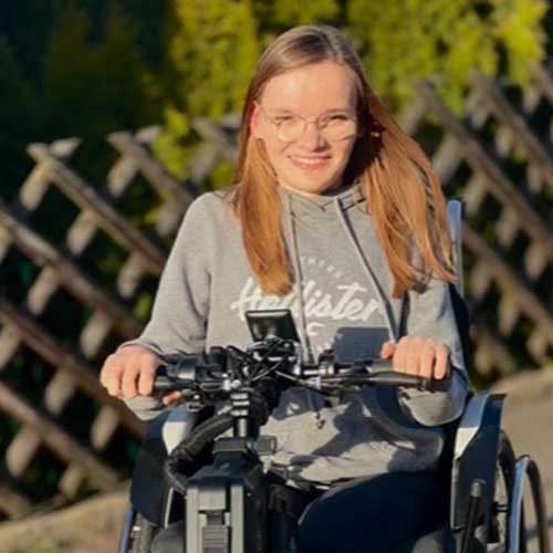 SMA-Betroffene Amelie fährt mit einem Zuggerät in ihrem Rollstuhl durch die Straßen und lächelt glücklich.