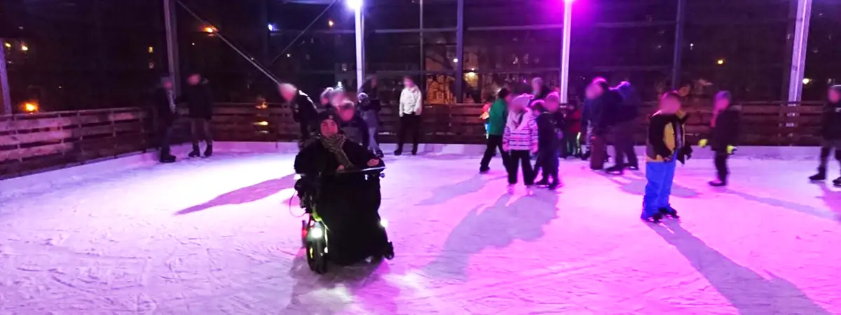 Joshua im Elektrorollstuhl auf dem Eis. Die Eisbahn ist mit lila Licht beleuchtet und im Hintergrund befinden sich noch andere Personen auf dem Eis. 