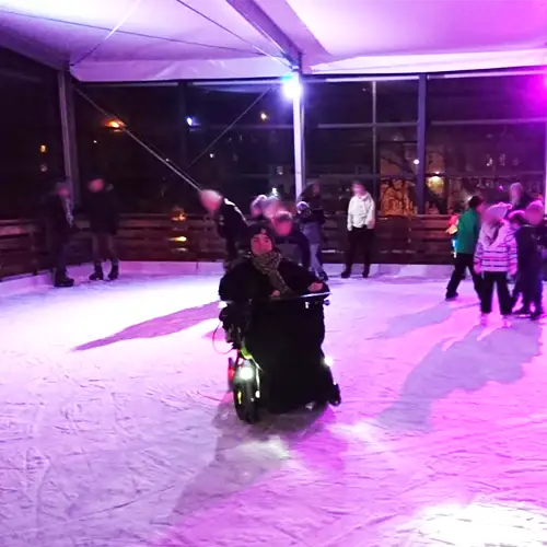 Joshua im Elektrorollstuhl auf dem Eis. Die Eisbahn ist mit lila Licht beleuchtet und im Hintergrund befinden sich noch andere Personen auf dem Eis. 