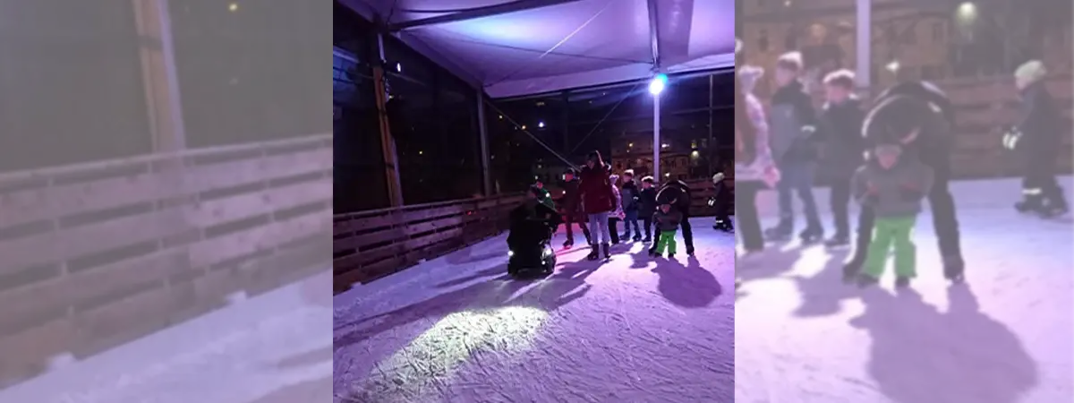 Joshua im Elektrorollstuhl auf dem Eis. Die Eisbahn ist mit blauem Licht beleuchtet und im Hintergrund befinden sich noch andere Personen auf dem Eis.