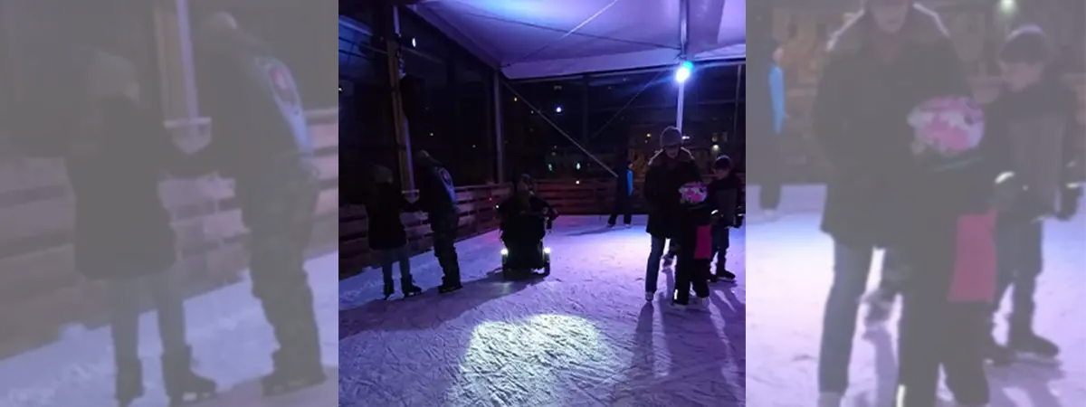 Joshua im Elektrorollstuhl auf dem Eis von weiter weg. Die Eisbahn ist mit lila Licht beleuchtet und im Hintergrund befinden sich noch andere Personen auf dem Eis.