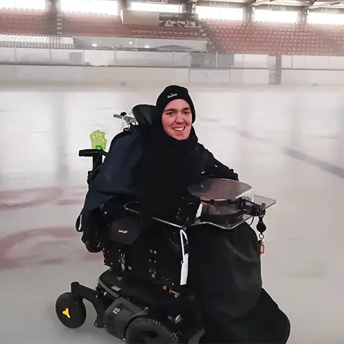 SMA-Betroffener Joshua sitzt in seinem Elektrorollstuhl auf einer Eislaufbahn. Er ist schwarz gekleidet und lächelt.