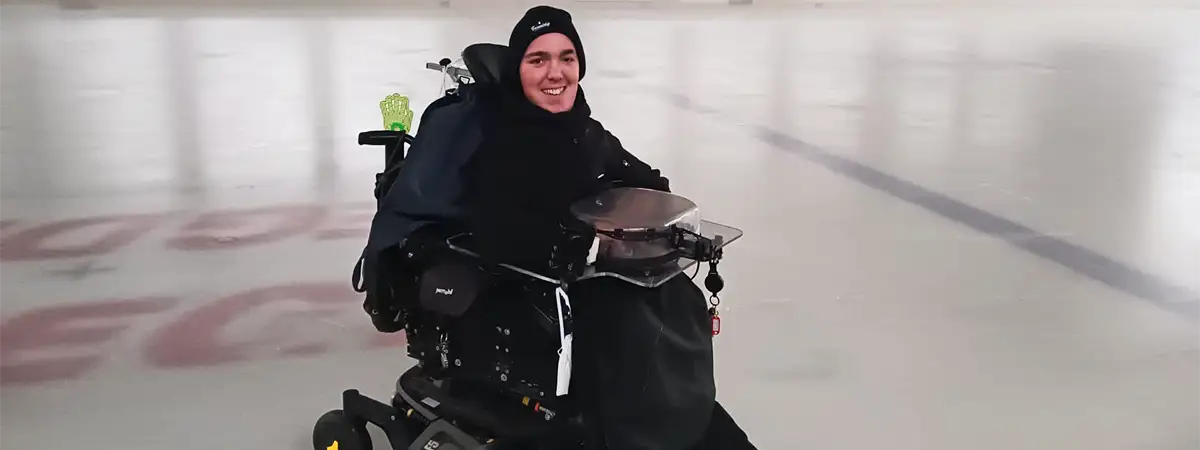 SMA-Betroffener Joshua sitzt in seinem Elektrorollstuhl auf einer Eislaufbahn. Er ist schwarz gekleidet und lächelt.
