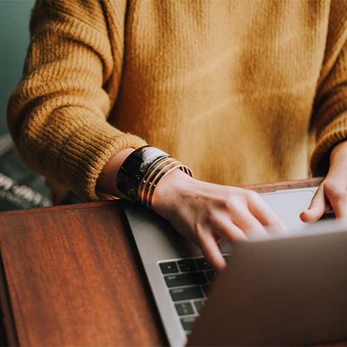 Eine Frau mit gelbem Pulli schreibt auf der Tastatur eines Laptops. 