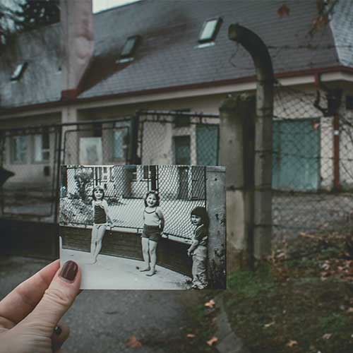 Ein altes Foto von drei Kindern, die vor einem Zaun an einer Schule stehen wird von einer Hand gehalten. Im Hintergrund sieht man das Gebäude des Fotos in seinem aktuellen, heruntergekommenen Zustand.