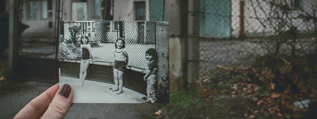 Ein altes Foto von drei Kindern, die vor einem Zaun an einer Schule stehen wird von einer Hand gehalten. Im Hintergrund sieht man das Gebäude des Fotos in seinem aktuellen, heruntergekommenen Zustand.