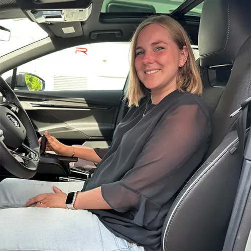 Foto von der Fahrerseite aus in den vorderen Innenraum des Autors der Autorin, die auf dem Fahrersitz sitzt mit Hand am Lenkrad. Sie trägt eine schwarze Bluse und eine hellblaue Jeans und schaut nach links aus dem Auto heraus.