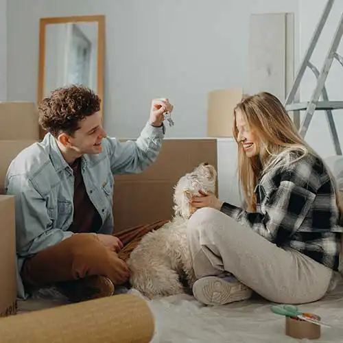 Ein junges Pärchen sitzt mit einem Hund auf dem Boden einer Wohnung, in der Umzugskartons stehen.