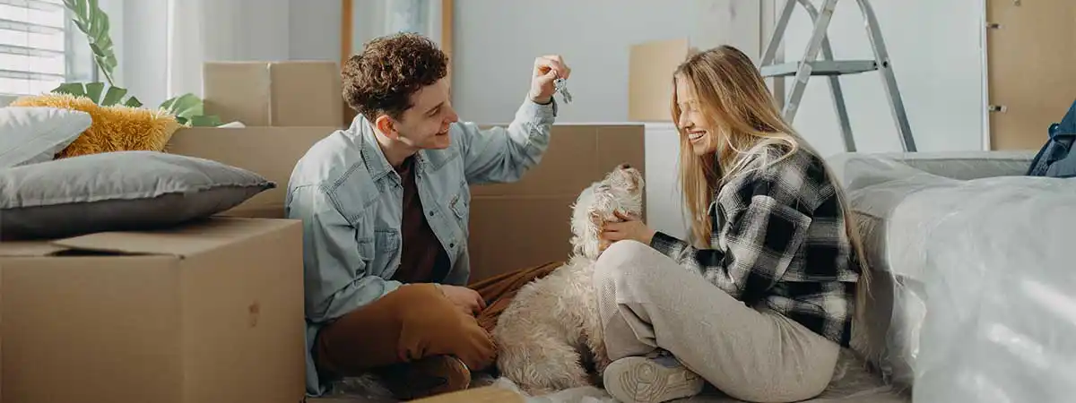 Ein junges Pärchen sitzt mit einem Hund auf dem Boden einer Wohnung, in der Umzugskartons stehen.