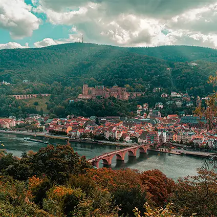 Ein Foto von der Stadt Heidelberg mit Fluss und Brücke, Häusern, Hügeln und Heidelberger Schloss. Sonnenstrahlen sind zu sehen.