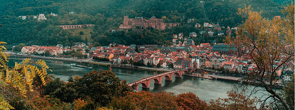 Ein Foto von der Stadt Heidelberg mit Fluss und Brücke, Häusern, Hügeln und Heidelberger Schloss. Sonnenstrahlen sind zu sehen.