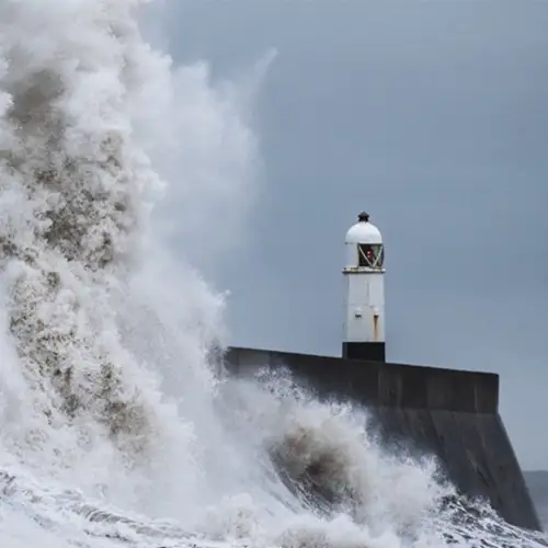 Ein Sturm an einer Küste. Grauer Himmel, Leuchtturm, Wellen, Wassermassen werden hochgeweht.