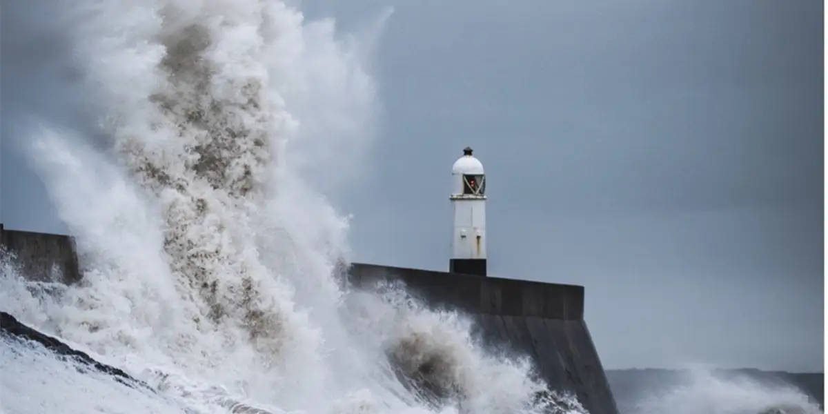 Ein Sturm an einer Küste. Grauer Himmel, Leuchtturm, Wellen, Wassermassen werden hochgeweht.