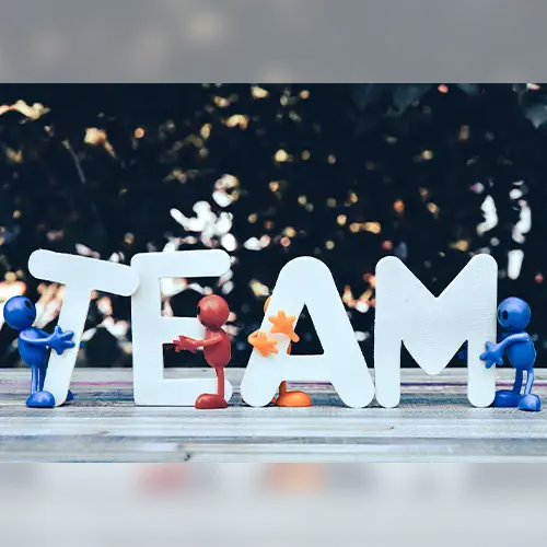 Foto von Buchstaben, die das Wort „Team“ bilden, auf einem Tisch stehen und die einzelnen Buchstaben werden von kleinen bunten Figuren umarmt. 
