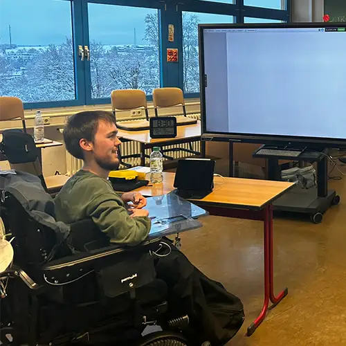 Foto in ein Klassenzimmer, in dem ein paar Schultische und Stühle zu sehen sind, sowie zwei große Bildschirme. Der Autor sitzt im Rollstuhl mit Blickrichtung zu den beiden großen Bildschirmen. 
