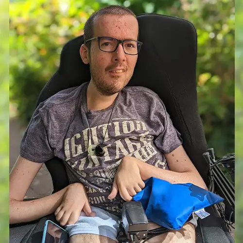 Foto des Autoren Roberto, der in seinem Rollstuhl sitzt und ein T-Shirt und eine kurze Hose trägt. Im Hintergrund ist ein grüner Busch zu sehen und die Sonne scheint. 