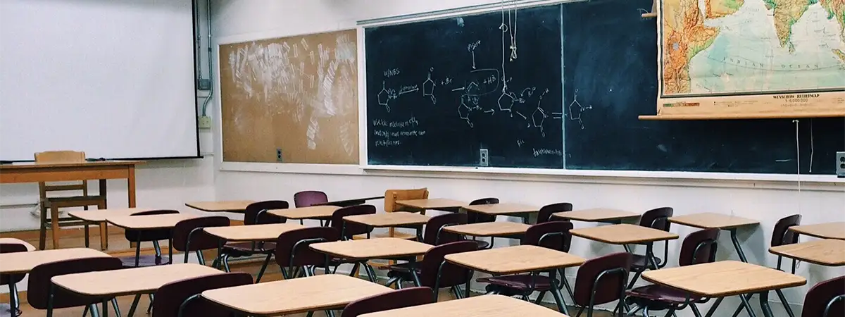 Aufnahme in einem Klassenzimmer mit mehreren einzelnen Pulten mit Stühlen, die nach vorne zum Lehrertisch zeigen. An der Rückwand sind mehrere Tafeln mit Kreidezeichnungen angebracht, von der Decke hängt eine Weltkarte.