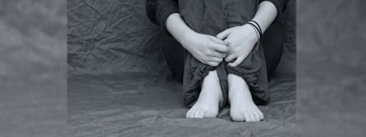 Ein schwarz-weißes Foto einer Person, die auf dem Boden sitzt und ihre Beine mit ihren Armen umschließt. Sie ist barfuß und nur der Unterkörper ist zu sehen.
