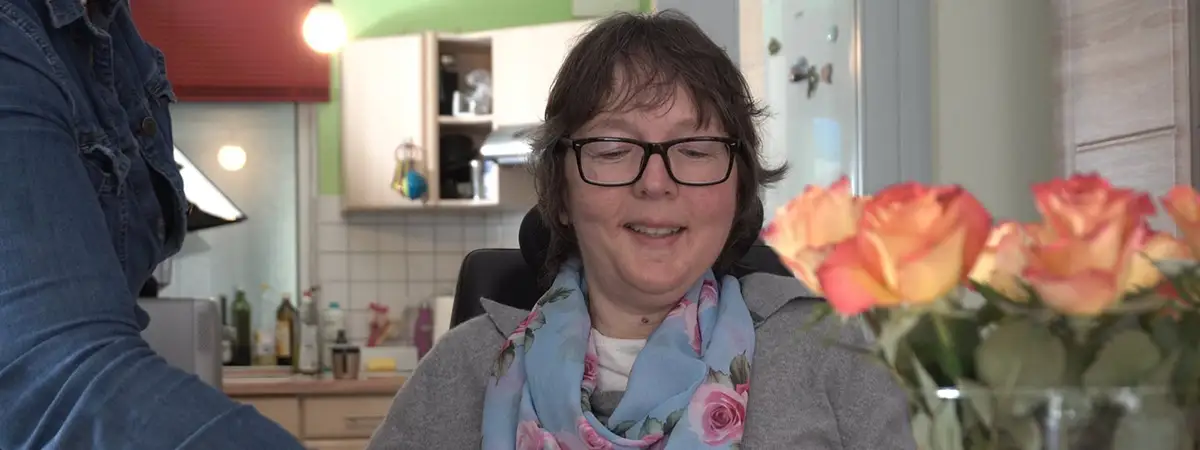 Ein Bild von Camilla, wie sie in ihrer Küche sitzt und lächelnd nach unten schaut. Im Vordergrund befinden sich orangefarbene Rosen und der Arm ihrer Assistenz, der seitlich ins Bild ragt.