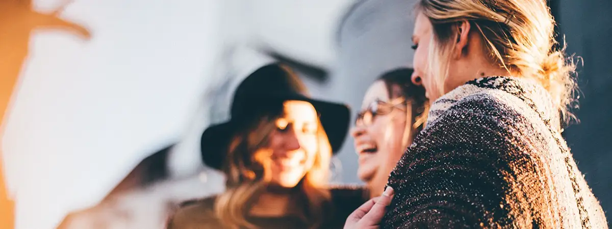 Drei Frauen stehen draußen in einer warmen, sonnigen Umgebung, lächeln und unterhalten sich miteinander. 