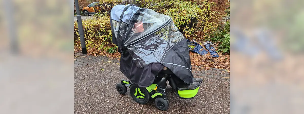 Ein Bild von Roberto, wie er in seinem elektrischen Rollstuhl sitzt, geschützt durch eine transparente Regenabdeckung, auf einem nassen Gehweg vor Büschen und Bäumen.