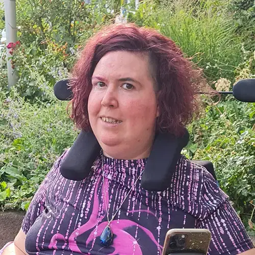 Ein Foto von Jasmin in ihrem elektrischen Rollstuhl. Sie trägt ein lilafarbenes T-Shirt und lächelt in die Kamera.