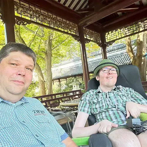 Roberto sitzt im Rollstuhl und trägt einen grünen Hut, eine kurze Hose und ein kurzärmeliges Karohemd. Neben ihm sitzt sein Partner in einem blau karierten Hemd. Beide schauen in die Kamera und befinden sich im Freien. Im Hintergrund sind grüne Bäume zu sehen und die Sonne scheint.