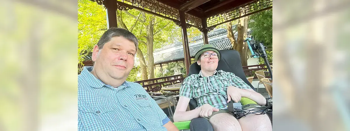 Roberto sitzt im Rollstuhl und trägt einen grünen Hut, eine kurze Hose und ein kurzärmeliges Karohemd. Neben ihm sitzt sein Partner in einem blau karierten Hemd. Beide schauen in die Kamera und befinden sich im Freien. Im Hintergrund sind grüne Bäume zu sehen und die Sonne scheint.