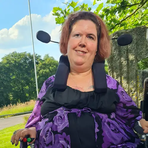 Ein Foto von Jasmin im Freien, die in ihrem Rollstuhl sitzt und freundlich in die Kamera lächelt. Sie trägt ein lila-schwarzes Oberteil und eine lange, lilafarbene Hose. Vor ihr liegt ihr selbstgeschriebenes Buch auf einem Tisch. 