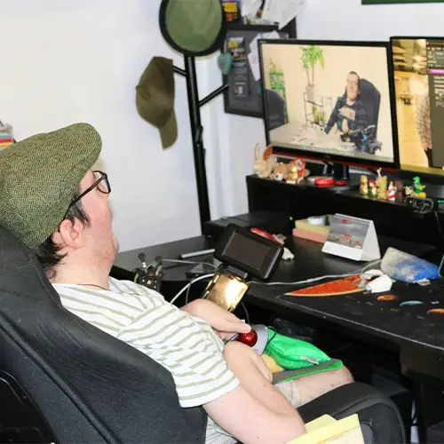 Ein Foto von Roberto in seinem Rollstuhl an einem Schreibtisch mit zwei Monitoren, die Text und ein Video zeigen, umgeben von persönlichen Gegenständen und Arbeitsmaterialien.