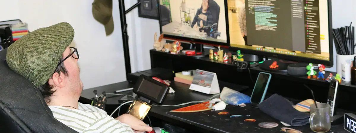 Ein Foto von Roberto in seinem Rollstuhl an einem Schreibtisch mit zwei Monitoren, die Text und ein Video zeigen, umgeben von persönlichen Gegenständen und Arbeitsmaterialien.